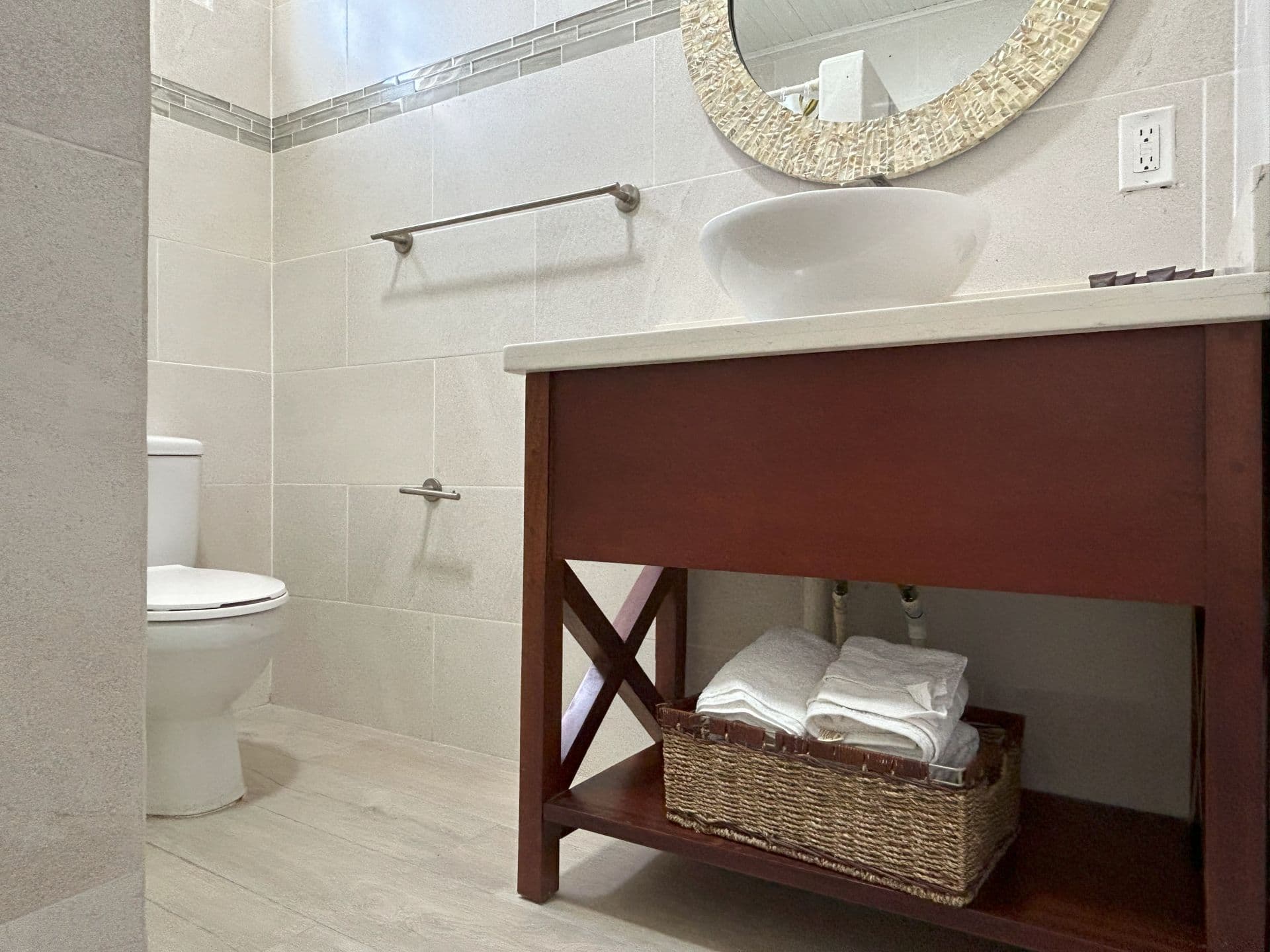 A brightly lit bathroom featuring a dark wood vanity with a vessel sink, a toilet, a decorative mirror, and light-tiled walls with an accent border.