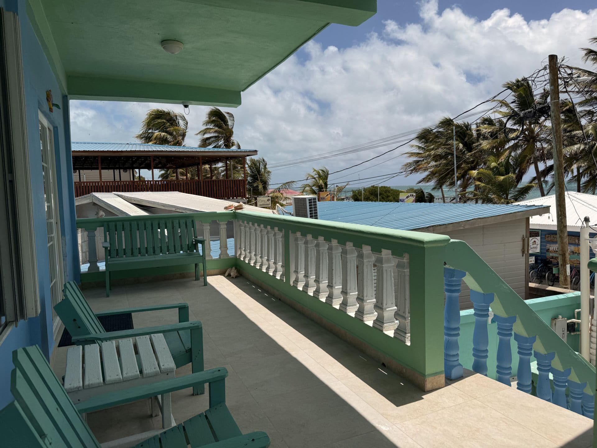 A vibrant turquoise and blue balcony with green-painted railings, offering a glimpse of distant palm trees and the ocean under a cloudy sky.