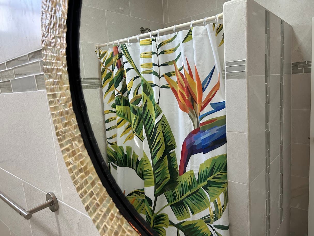 A tiled bathroom featuring a shower with a tropical-themed shower curtain with bird of paradise flowers and lush leaves reflected in a stylish mirror.
