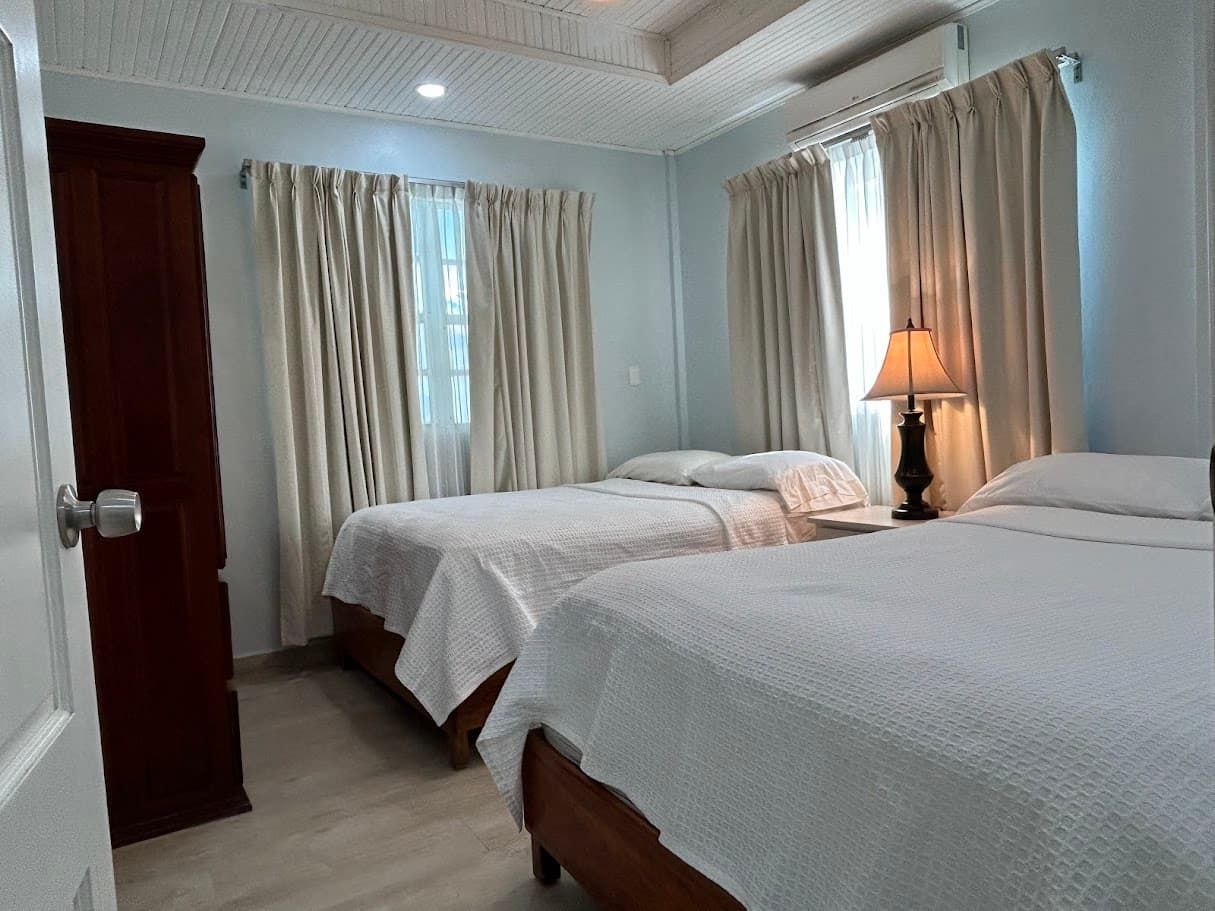 A well-lit bedroom featuring two comfortable beds with white bedding, a wooden wardrobe, a bedside lamp, and two windows with neutral curtains.