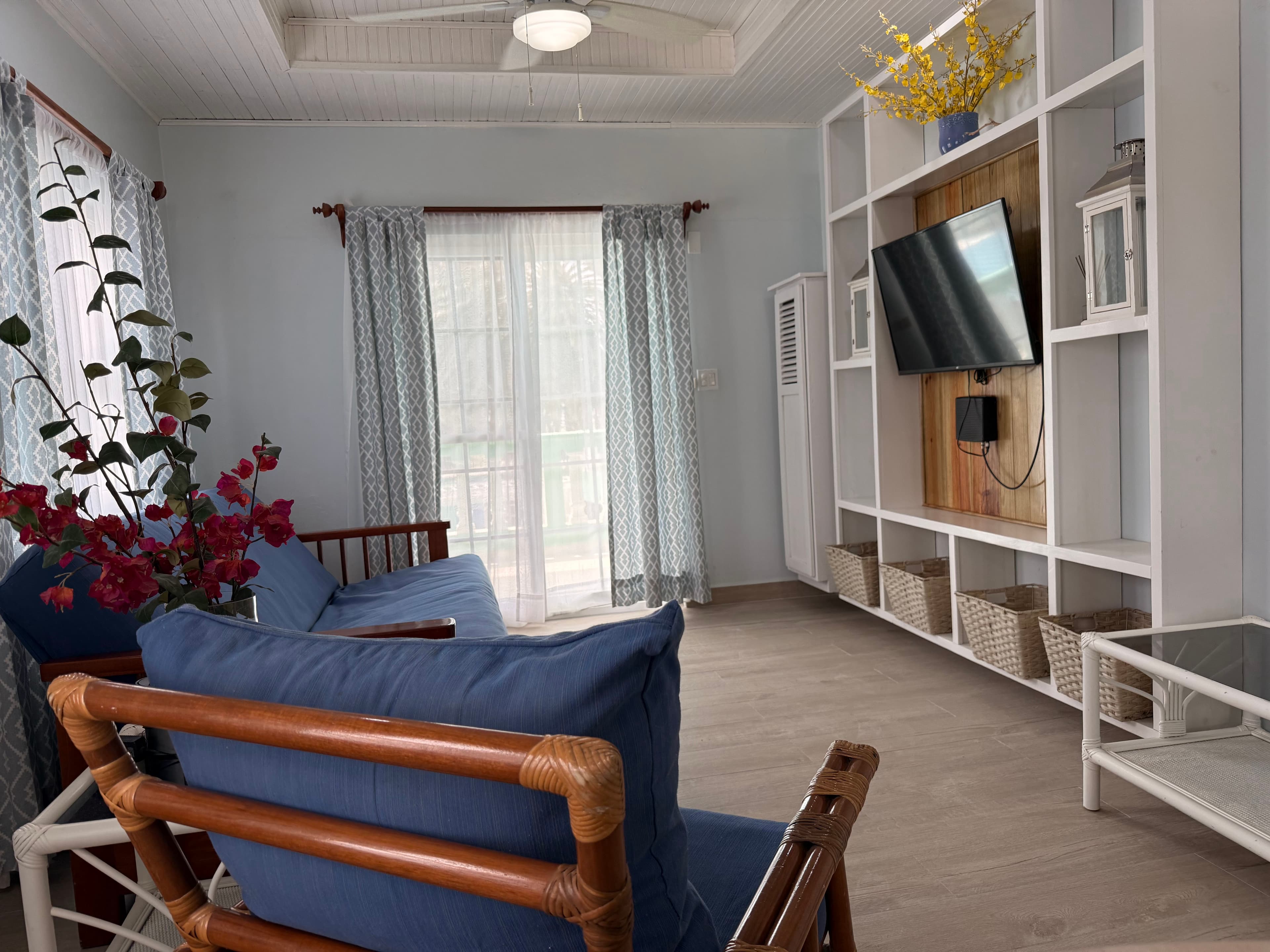 A modern living space featuring a blue sofa and armchair, a large entertainment unit with a mounted TV, and a bright window with sheer curtains.