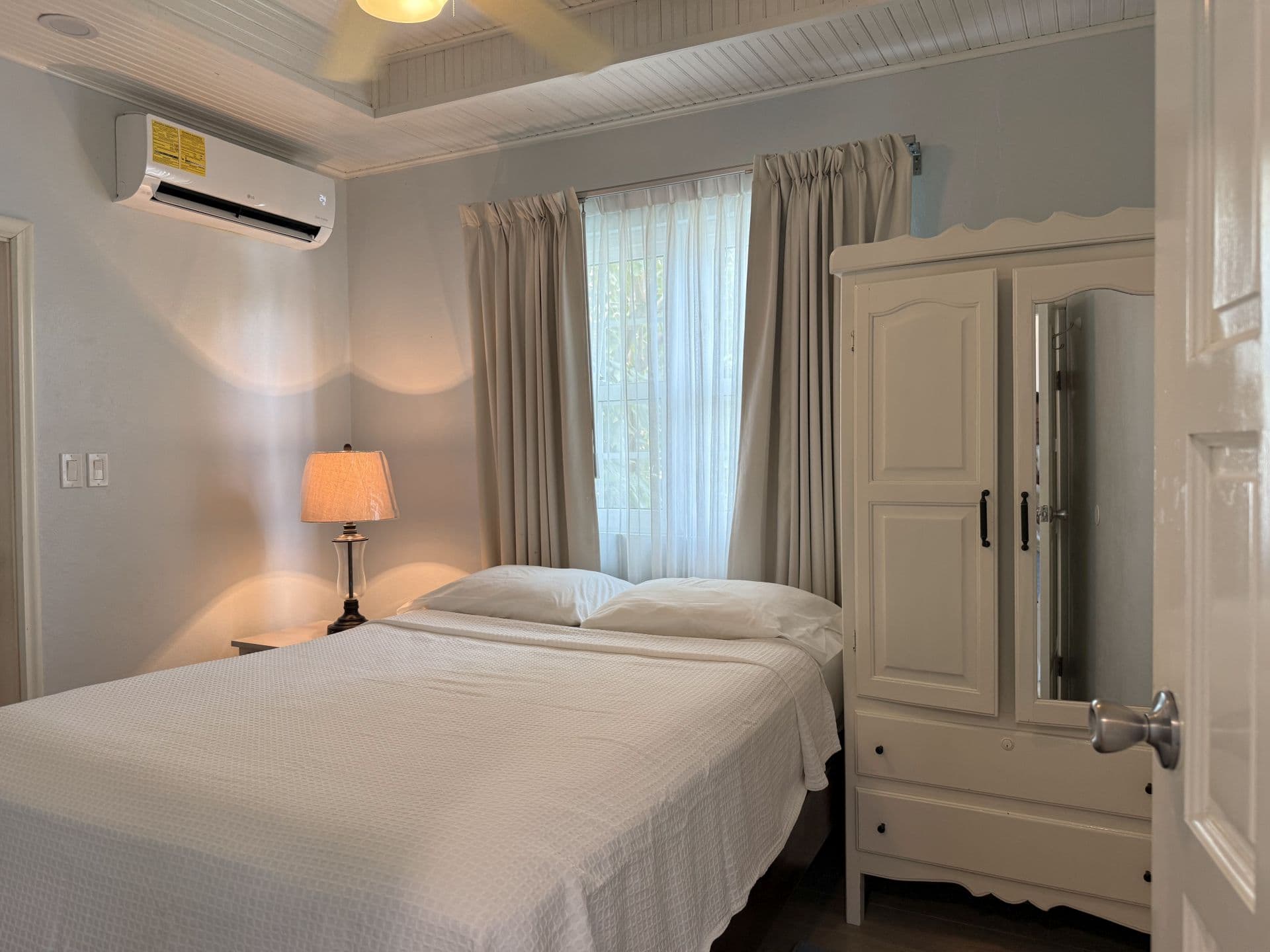 A serene bedroom featuring a comfortable bed with white linens, a classic white wardrobe with a mirrored door, and a window with neutral curtains.