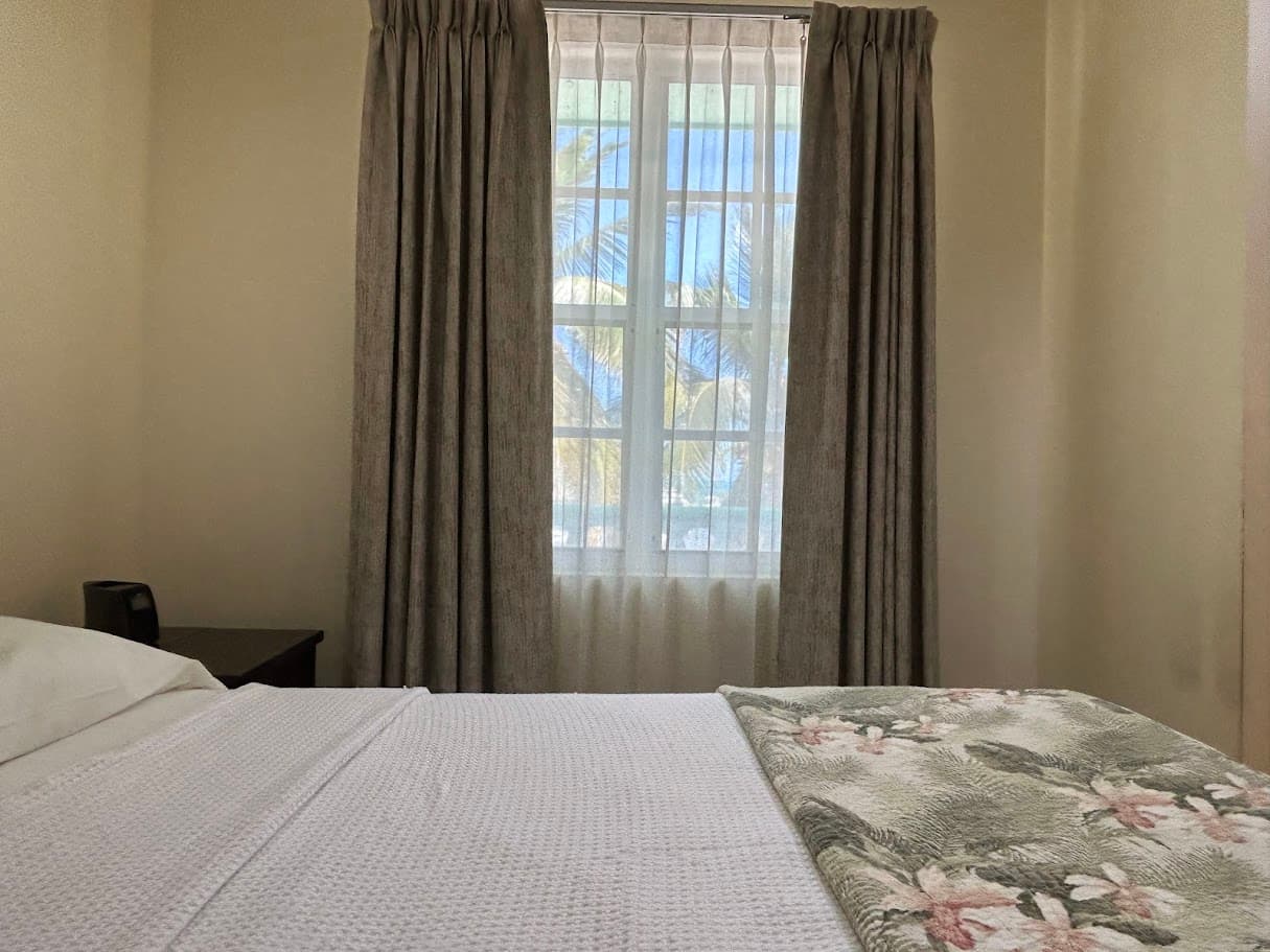 A serene bedroom scene highlighting a bed with a floral throw, beside a large window framed by beige curtains and sheer drapes.