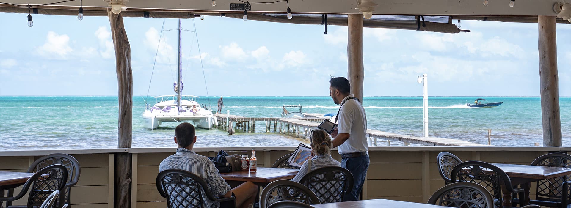 A view from a seaside restaurant featuring patrons at tables, a dock with a boat, and turquoise waters beyond.