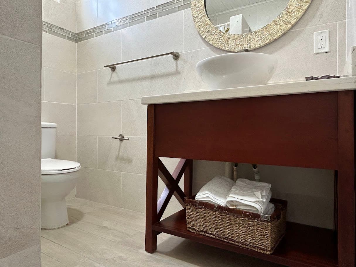 A bright bathroom featuring a dark wood vanity with a vessel sink, a toilet, a decorative mirror, and light-tiled walls with an accent border.