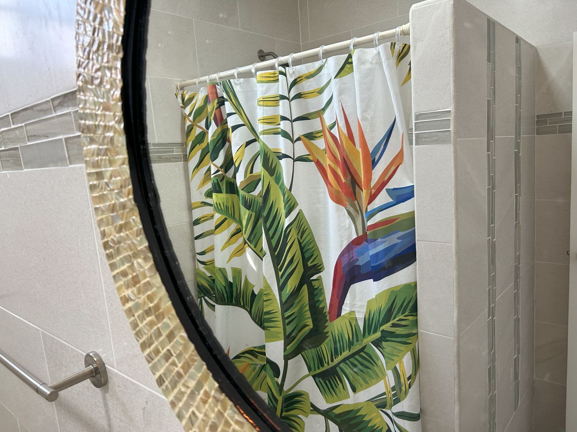 A tiled bathroom featuring a shower with a tropical-themed shower curtain with bird of paradise flowers and lush leaves reflected in a stylish mirror.
