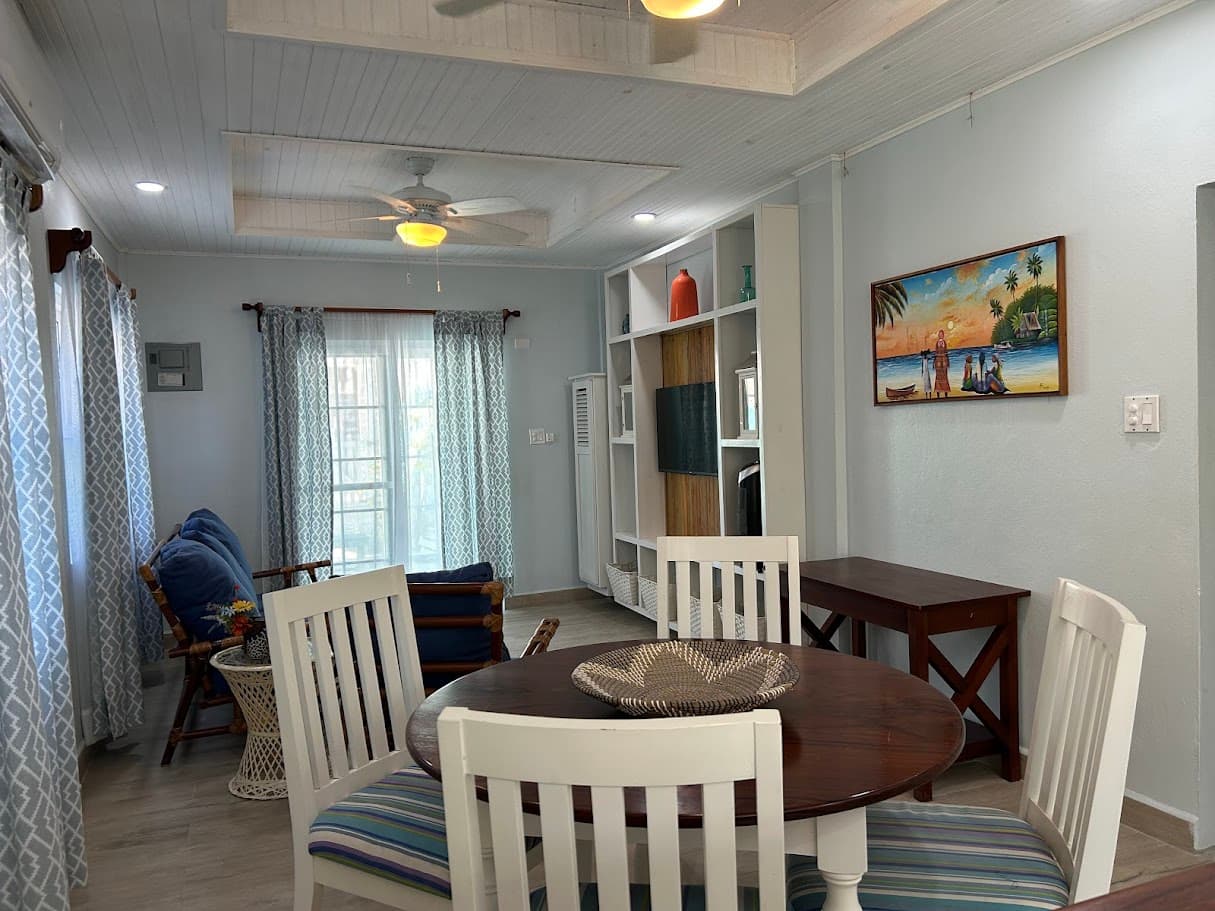 A brightly lit living and dining area with a round wooden table, white chairs, a comfortable sofa, and a wall-mounted TV cabinet.
