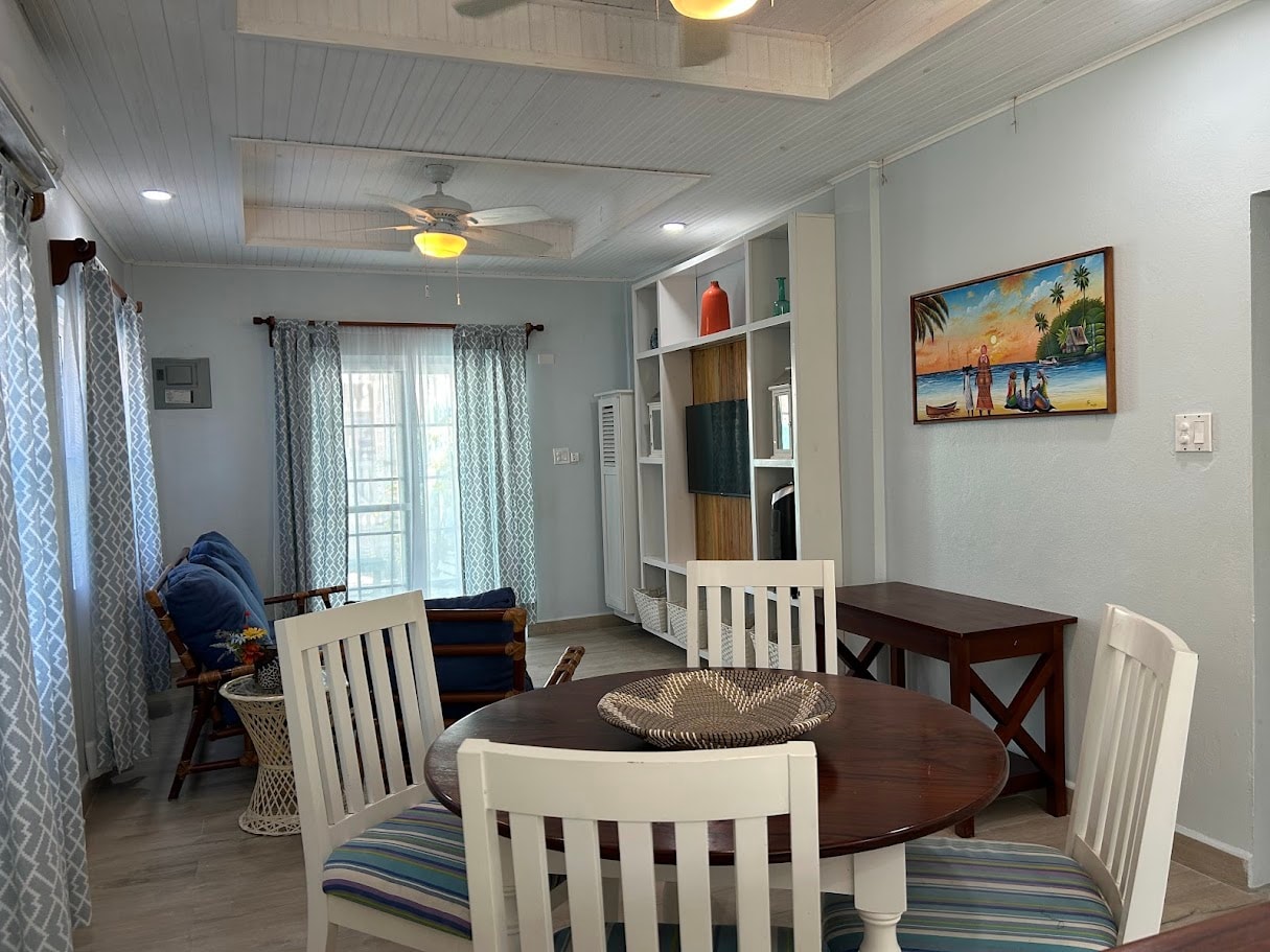 A brightly lit living and dining area with a round wooden table, white chairs, a comfortable sofa, and a wall-mounted TV cabinet.