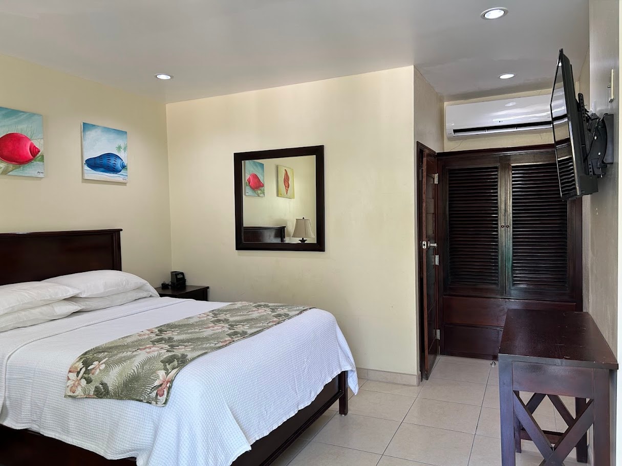 A hotel room featuring a queen-size bed with a floral blanket, tropical fish artwork, a mirror, and a wall-mounted TV.