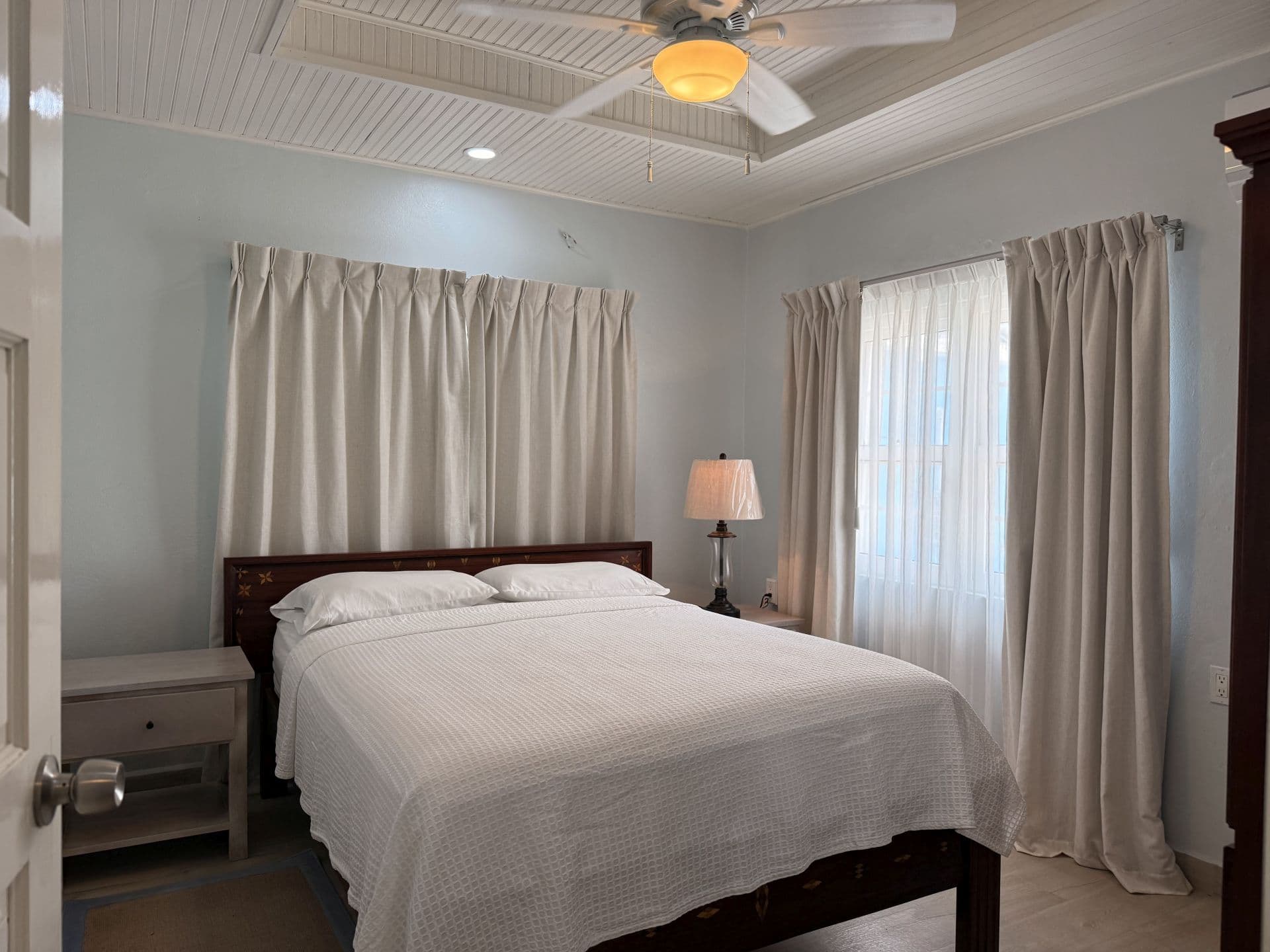 A well-lit bedroom featuring a queen-sized bed with white linens, a bedside table with a lamp, a ceiling fan, and large windows with cream-colored curtains.