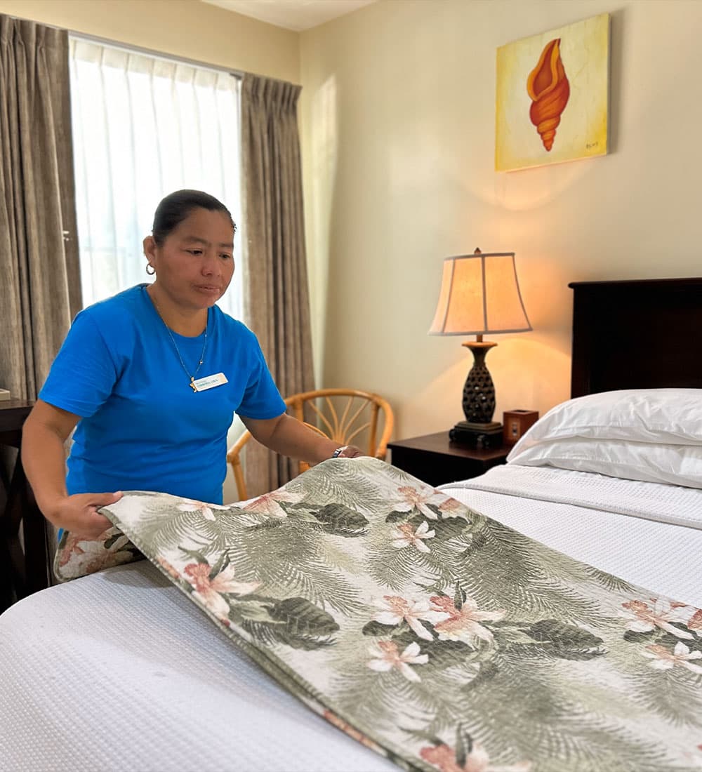 A hotel staff member in a blue shirt arranges a floral blanket on a made bed in a well-lit room.