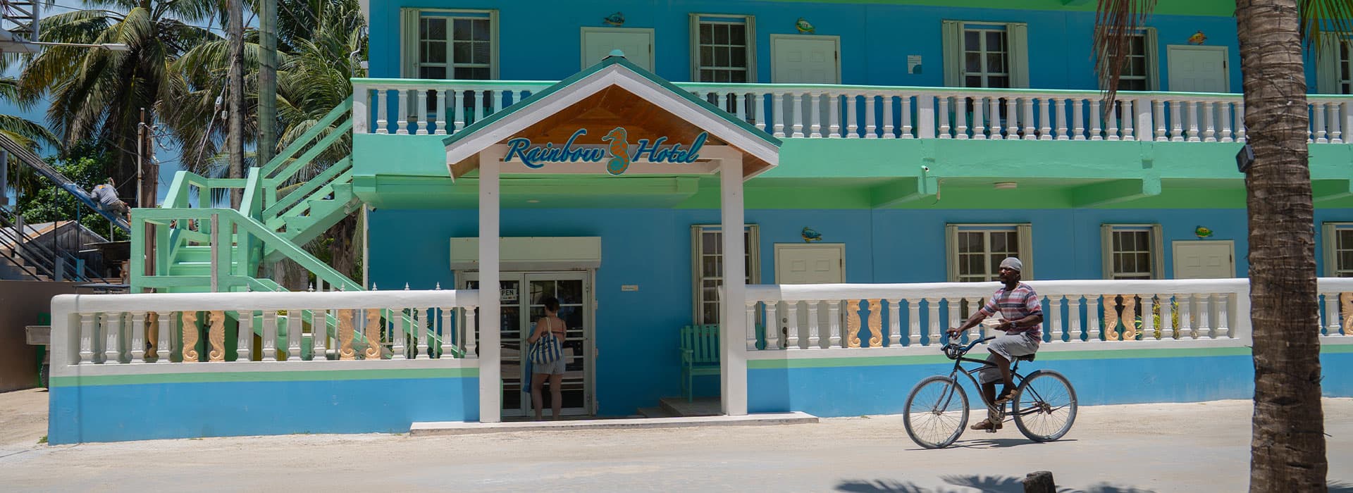 A man rides a bicycle past the colorful entrance of the Rainbow Hotel surrounded by palm trees.
