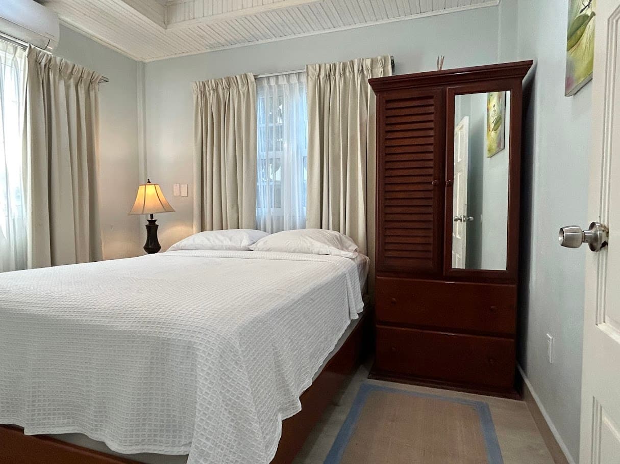 A tranquil bedroom with a comfortable bed, a classic wooden wardrobe with a mirror, a bedside lamp, and a window with light-colored curtains.