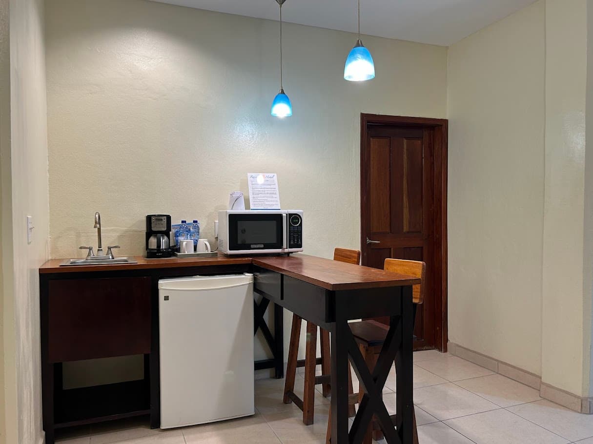 A compact kitchenette area with a microwave, coffee maker, mini-fridge, sink, and a high-top breakfast bar with stools, under blue pendant lights.