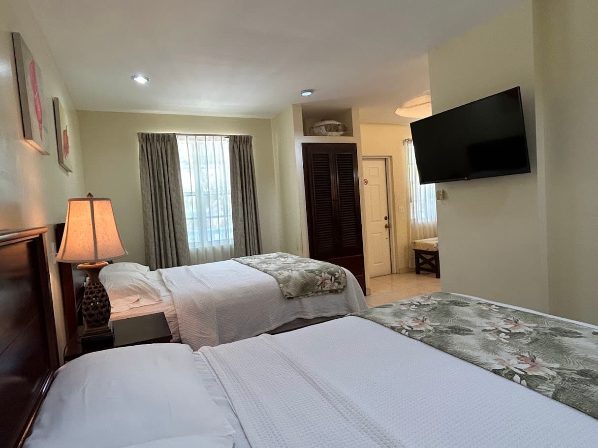 A spacious hotel room with two beds, each adorned with a floral throw, a bedside lamp, a flat-screen TV, and a large window with sheer curtains.